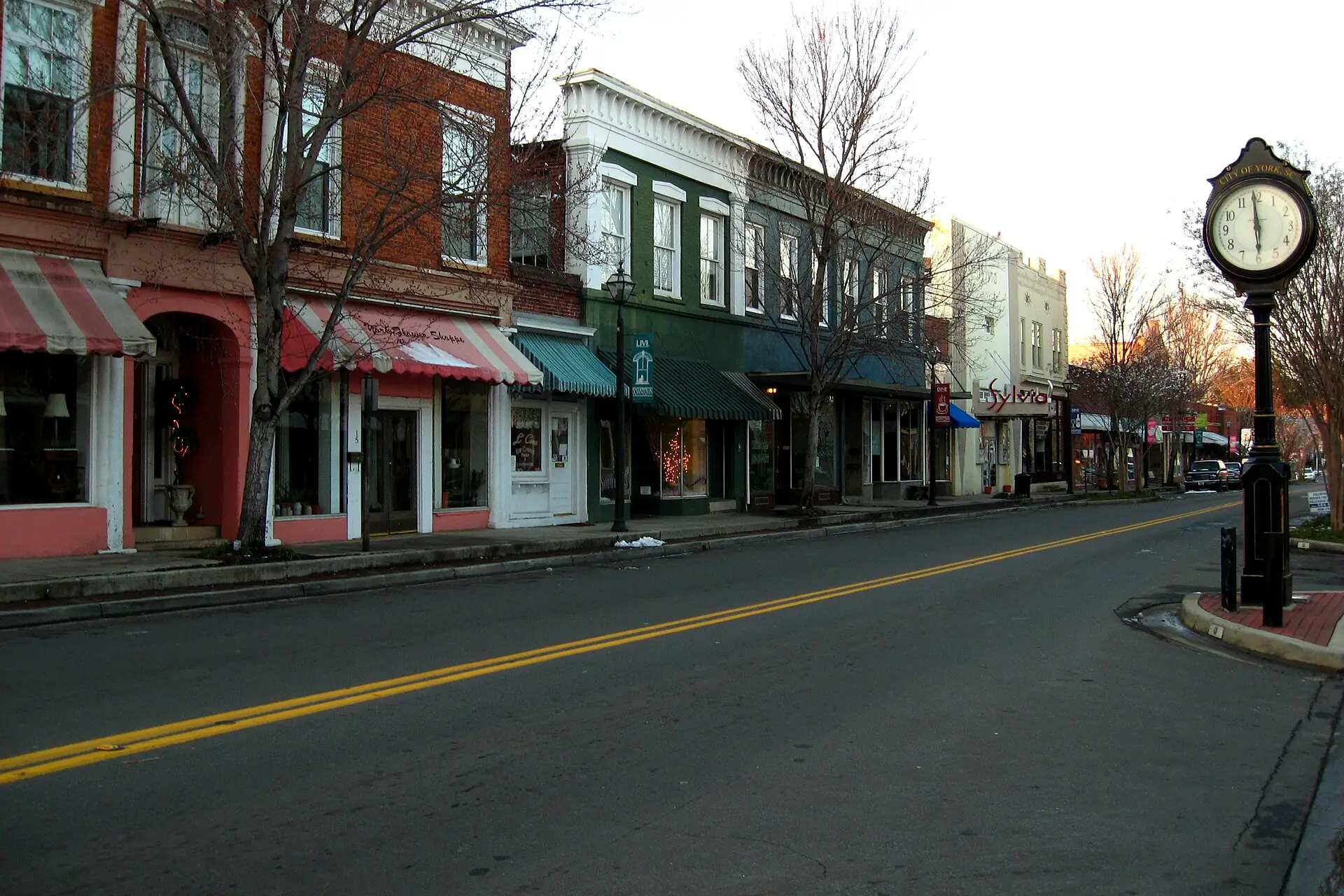 Brick storefronts with striped awnings along a street in downtown York, SC, beside a City of York street clock